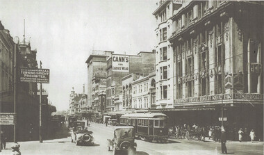 A busy city street with vintage cars and trams, flanked by multi-story buildings with shops and signs including "Cann's for Ladies Wear" and "Square Deal Sale Now On".