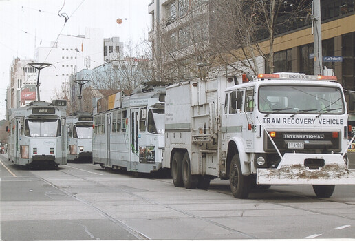 A white tram recovery vehicle with "TRAM RECOVERY VEHICLE INTERNATIONAL" written on its side tows two vintage grey trams down a city street with buildings and overhead wires visible.