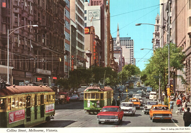 Vintage view of a busy city street with trams and cars, featuring tall buildings and trees under a clear blue sky.