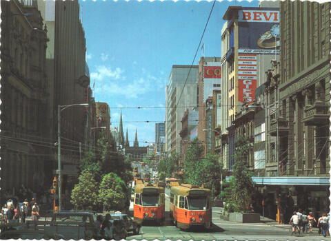 Vintage view of a wide city street lined with tall buildings, featuring two orange and yellow trams on tracks, with pedestrians and trees.
