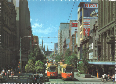 Vintage view of a wide city street lined with tall buildings, featuring two orange and yellow trams on tracks, with pedestrians and trees.