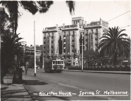 A vintage tram on a street in front of Alcaston House on Spring Street, Melbourne, with palm trees in the foreground.