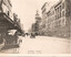 A bustling Bourke Street in Melbourne, Victoria, featuring classic automobiles and pedestrians on a wide sidewalk lined with ornate 1920s architecture, including a prominent clock tower in the distance and one cable tram.