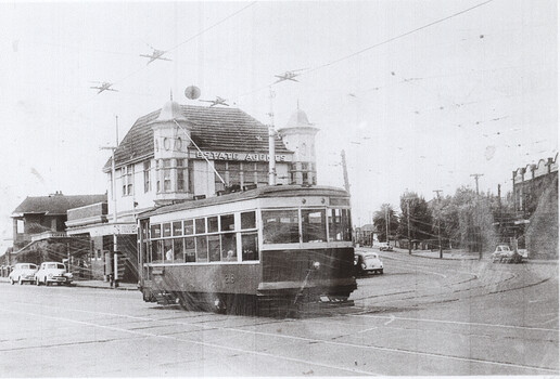 A vintage tram car pauses at a street intersection in front of a ornate brick building with towers, with several cars visible on the road.