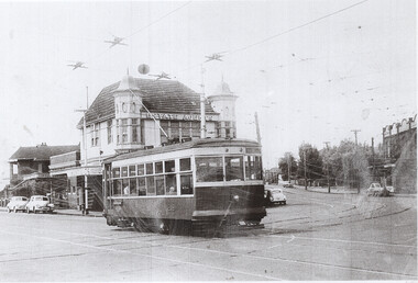 A vintage tram car pauses at a street intersection in front of a ornate brick building with towers, with several cars visible on the road.
