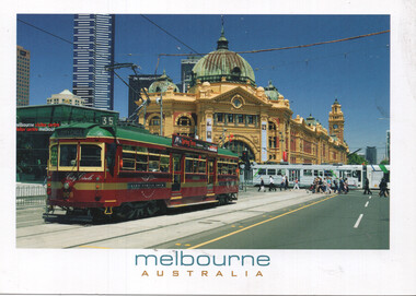 A vintage red and green tram travels along tracks in front of the grand yellow facade of Flinders Street Station in Melbourne, Australia, with modern skyscrapers in the background and people crossing a street in the distance.