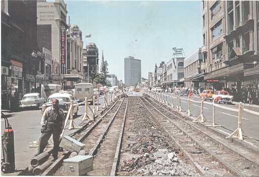Street scene with former cable tram tracks being removed, flanked by wooden barriers and construction materials. Several vintage cars are visible on the right side of the street, with shops and tall buildings lining both sides. A worker stands on the left, next to pipes and a toolbox.