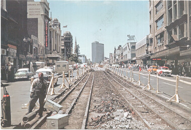 Street scene with former cable tram tracks being removed, flanked by wooden barriers and construction materials. Several vintage cars are visible on the right side of the street, with shops and tall buildings lining both sides. A worker stands on the left, next to pipes and a toolbox.