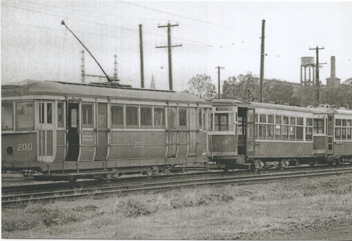 A line of vintage streetcars numbered 200, 208, and 217, parked on a railway track with industrial buildings and a water tower in the background.