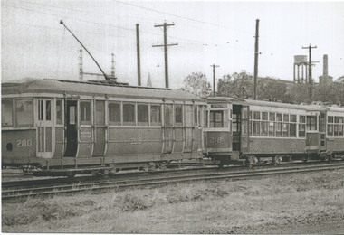 A line of vintage streetcars numbered 200, 208, and 217, parked on a railway track with industrial buildings and a water tower in the background.