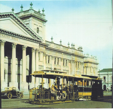 A yellow cable tramcar with passengers travels along a city street in front of an ornate neoclassical building with a grand colonnade.