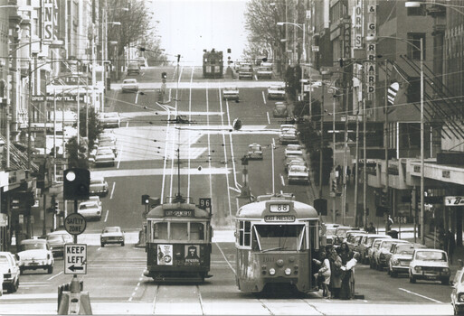 Two trams travel down a busy, wide city street lined with shops and parked cars under a grey sky.