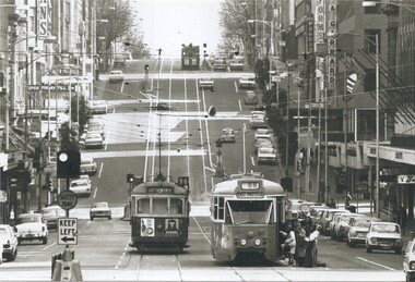 Two trams travel down a busy, wide city street lined with shops and parked cars under a grey sky.