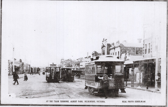 Early 20th-century cable trams lined up on a street with buildings and pedestrians.