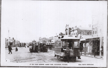 Early 20th-century cable trams lined up on a street with buildings and pedestrians.