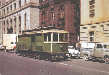 A vintage green tram with "Freight car" visible on its side travels down a city street lined with historic buildings. Several cars and a delivery truck are parked or moving nearby.