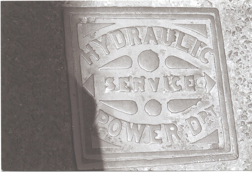 Close-up, overhead view of a square metal plate embedded in the ground, featuring embossed text reading "HYDRAULIC SERVICE POWER D.".