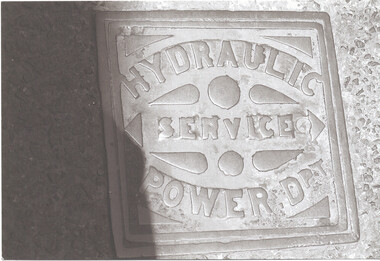 Close-up, overhead view of a square metal plate embedded in the ground, featuring embossed text reading "HYDRAULIC SERVICE POWER D.".