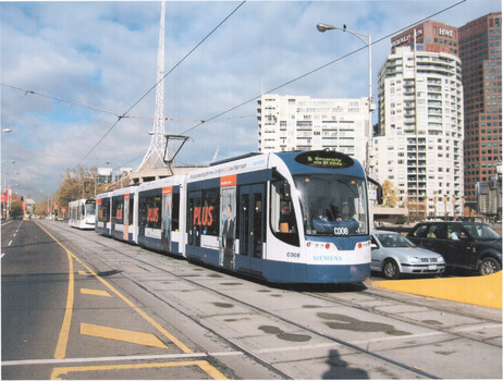 Modern blue and white Siemens tram with advertisements along its side, traveling on tracks on a city street next to cars, with tall buildings in the background under a cloudy sky.