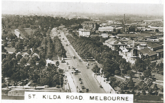 Aerial view of a wide, tree-lined boulevard with heavy traffic, leading towards a cityscape of buildings under a hazy sky, with the text "ST. KILDA ROAD. MELBOURNE" in the foreground.