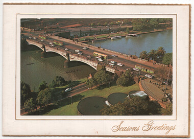 Aerial view of a wide, multi-lane bridge spanning a body of water, with traffic including cars and trams. Lush green trees and manicured lawns line the banks, with a section of a circular fountain visible in the foreground.