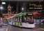 Modern green and white tram on city tracks at night, with a historic clock tower building in the background.