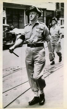 A soldier in a wide-brimmed slouch hat, khaki uniform with a dark armband, and boots walks with a baton in his outstretched hand down a street with tram tracks.