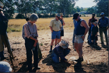 Group of approximately 10 people stand and watch a kneeling man set a trap
