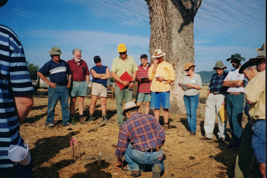 Colour photograph depicting twelve members of Wooragee Landcare Group or local community members watching a kneeling man attend to a trap. 