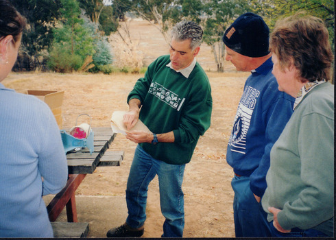 Three people stand outside beside a picnic table and watch a man as he points to a piece of paper in his hands.  