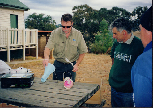 Three men stand outside around a picnic table observing two tube shaped objects. One man handles one of the tubes. 