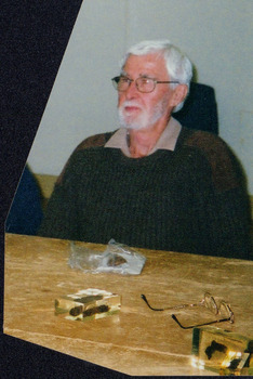 Image of man looking at a speaker (not pictured). On the table in front of him is two natural specimens in resin and a pair of spectacles. 