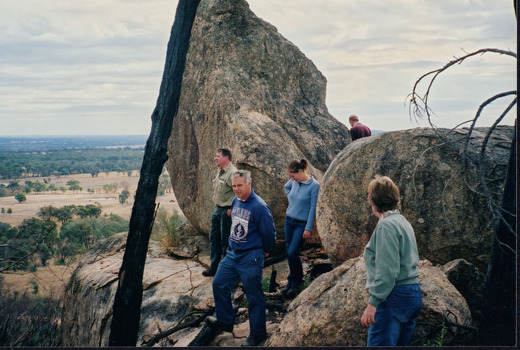 Photograph of three men and two women looking out over a cliff. They are surrounded by large boulders.