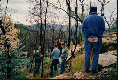 Colour photograph showing five people standing on the side of a sloping, rocky, wooded bushland. Four people are grouped together in middle background appearing to be in conversation, while one person is standing on a rock closer to the photographer facing the others with hands clasped together behind their back.with back to camera.
