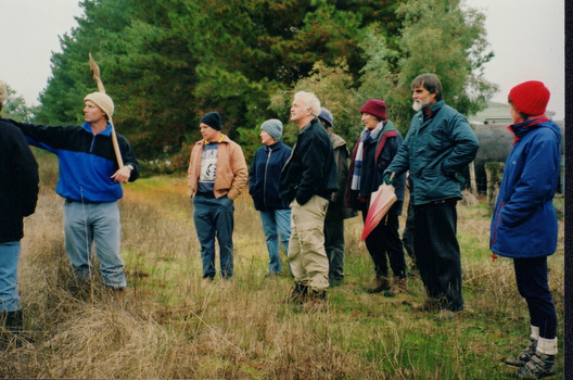 Colour photograph showing ten people standing in a paddock. One person on the left of the photograph has a gardening tool over their shoulder and appears to be pointing to and discussing something out of sight. 