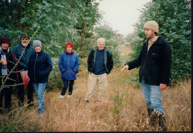 Colour photograph showing six people standing in a bushy native grassland or edge of a paddock. One person on the right of the photograph appears to be mid demonstration gesturing to a small bush in the grass.