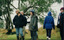Colour photograph of six people in the bushland, wearing winter clothes. Four of the group are looking left towards the distance, one person is looking directly at the photographer, and one is walking away with their back to the camera..