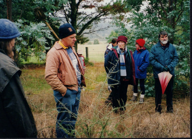 Colour photograph of six people in the bushland, attending the tree planting field day. Four of them are looking towards the same direction and two of them are talking to each other.