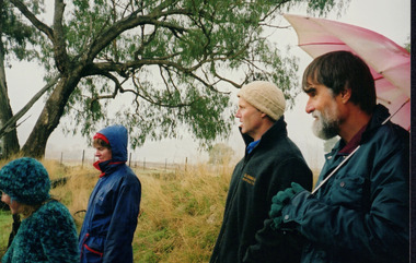 Coloured photograph of 4 individuals, two male and two female, in thick jackets, beanies with one man holding an umbrella. There is a grassy field in the background with an old fence and a large tree.