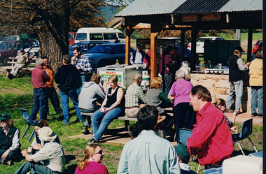 Coloured photograph of a park with 24 people visible in the shot. Some are sitting at a bench while others are sitting on the grass, some are standing up in groups and others are under the small outdoor kitchen area. The people within the photograph appear to be dressed casually.