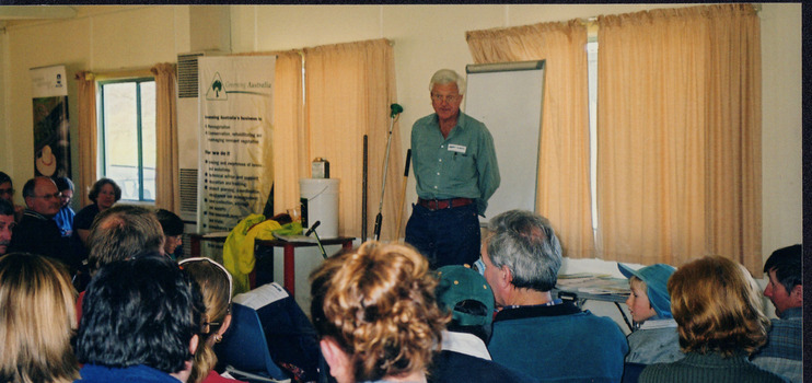 Group of people at a meeting in an indoor settling with an audience and speaker. The group includes men, women, and children.