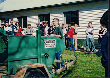 Group of people in an outdoor setting watching a demonstration by a male speaker in front of a harvester. The group includes men, women, and children.