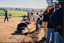 A large group of people stand and crouch over a newly dug-up winding line of dirt in the ground. 