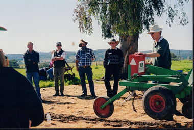 Four men stand side-by-side listening to a fifth man talking whilst placing his hand on a large piece of farming equipment. 