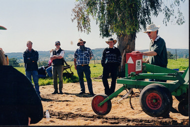 Four men stand side-by-side listening to a fifth man talking whilst placing his hand on a large piece of farming equipment. 