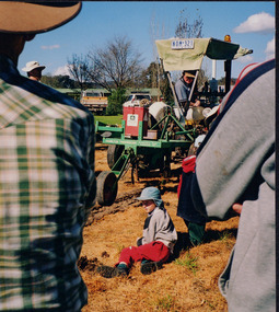 Photograph taken behind the backs of two onlookers observing a man working a medium-sized piece of farming equipment whilst a child sits on the ground beside it. 