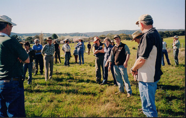 A group of people stand together in an open field, exchanging conversation. 