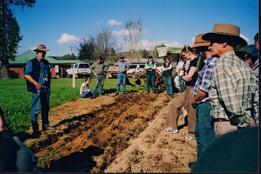 Colour photograph of ten people standing in a circle around a patch of dirt.