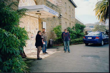 Three people standing outside an brick building with a blue car to the far right.