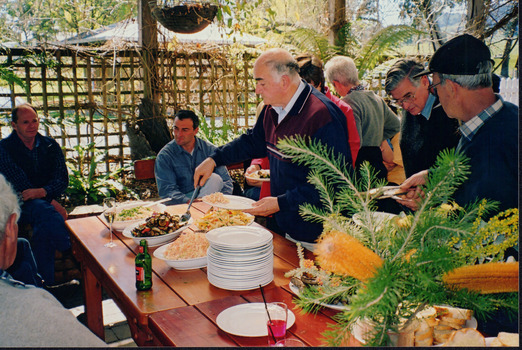 A man is serving himself food from a table in front of him. Other men appear to be waiting to also serve themselves food, with two others sitting in the back of the photograph.